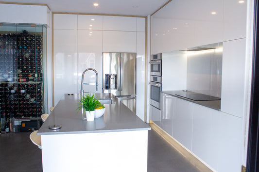 Cuisine moderne Vision Renov avec îlot central en quartz gris, armoires blanches, électroménagers en acier inoxydable, cave à vin vitrée et plaque de cuisson intégrée. Décorée avec une plante verte et un bol de fruits. Germoir-02.