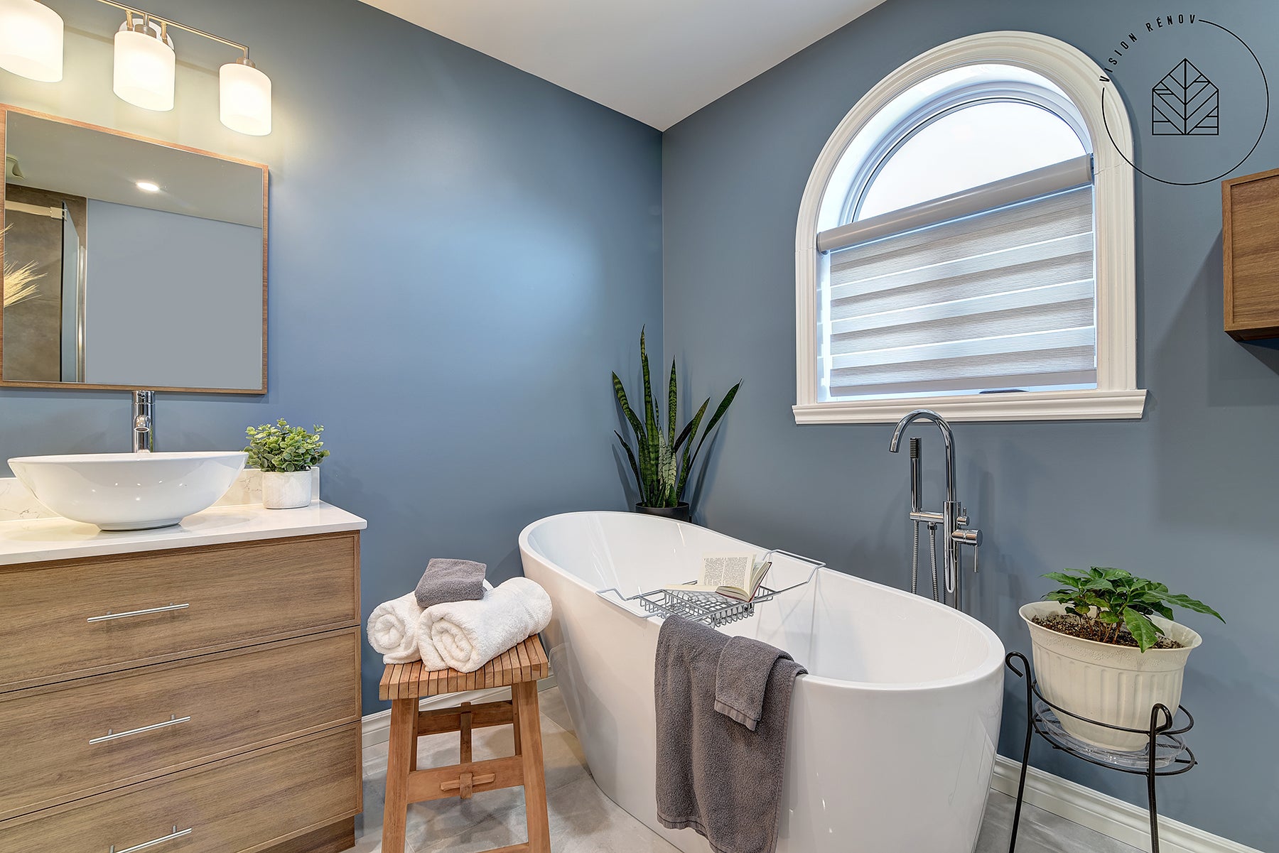 Salle de bain moderne avec baignoire autoportante blanche, murs bleu-gris, meuble-lavabo en bois, miroir encadré, et décoration avec plantes vertes et serviettes soigneusement disposées. Fenêtre arquée avec store tamisant la lumière naturelle.
