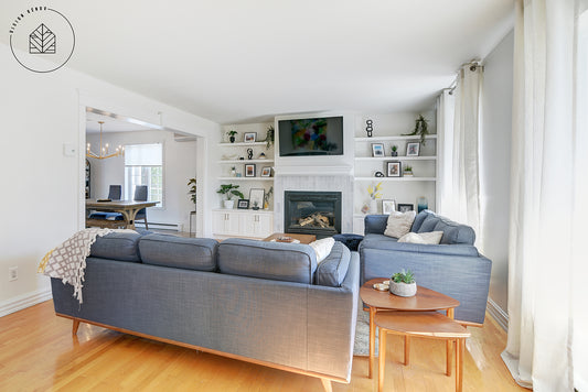 Salon moderne Vision Renov avec canapés gris, table basse en bois, cheminée blanche, TV murale, étagères décoratives et rideaux blancs. Ouverture vers une salle à manger avec table et lustre. Ritournelle-01.